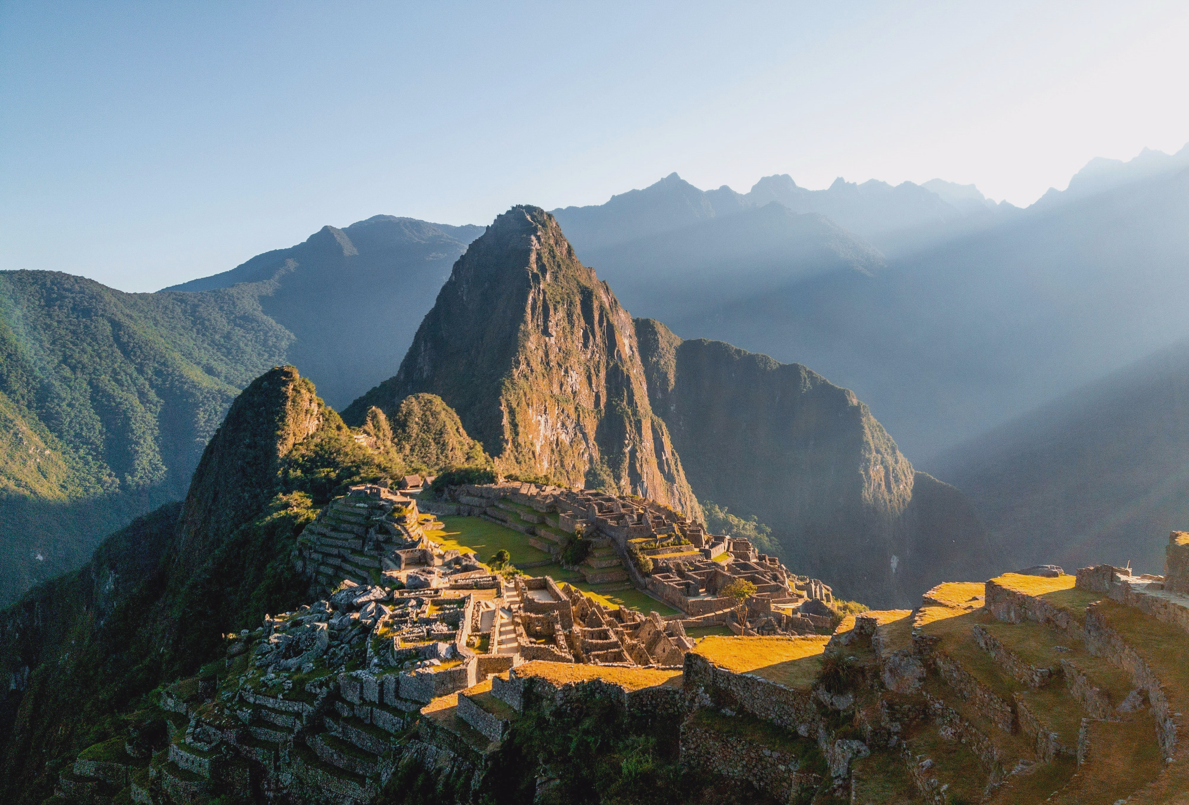 Machu Picchu's stone buildings and green terraces spread below Huayna Picchu in warm morning light.