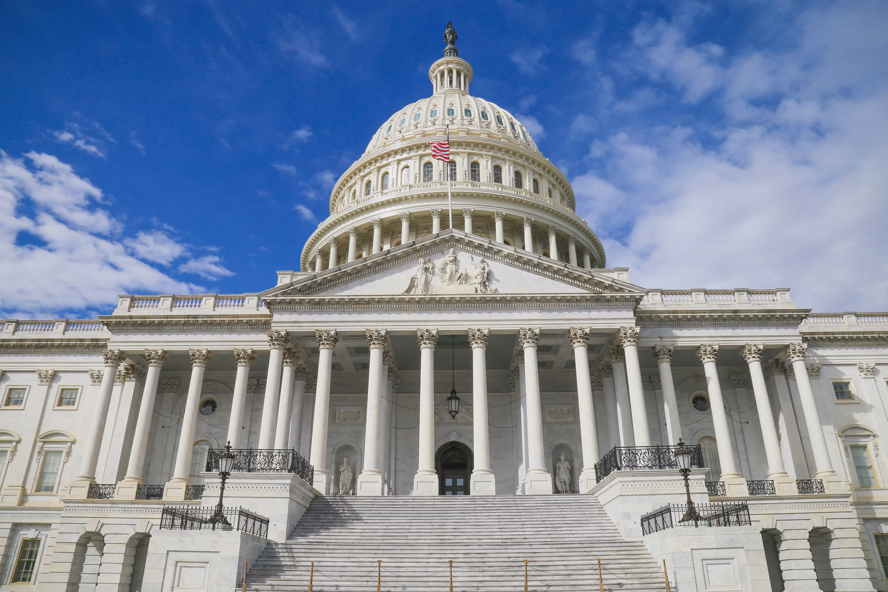 Low-angle view of the U.S. Capitol's white colonnade and dome beneath a bright blue, cloud-filled sky.