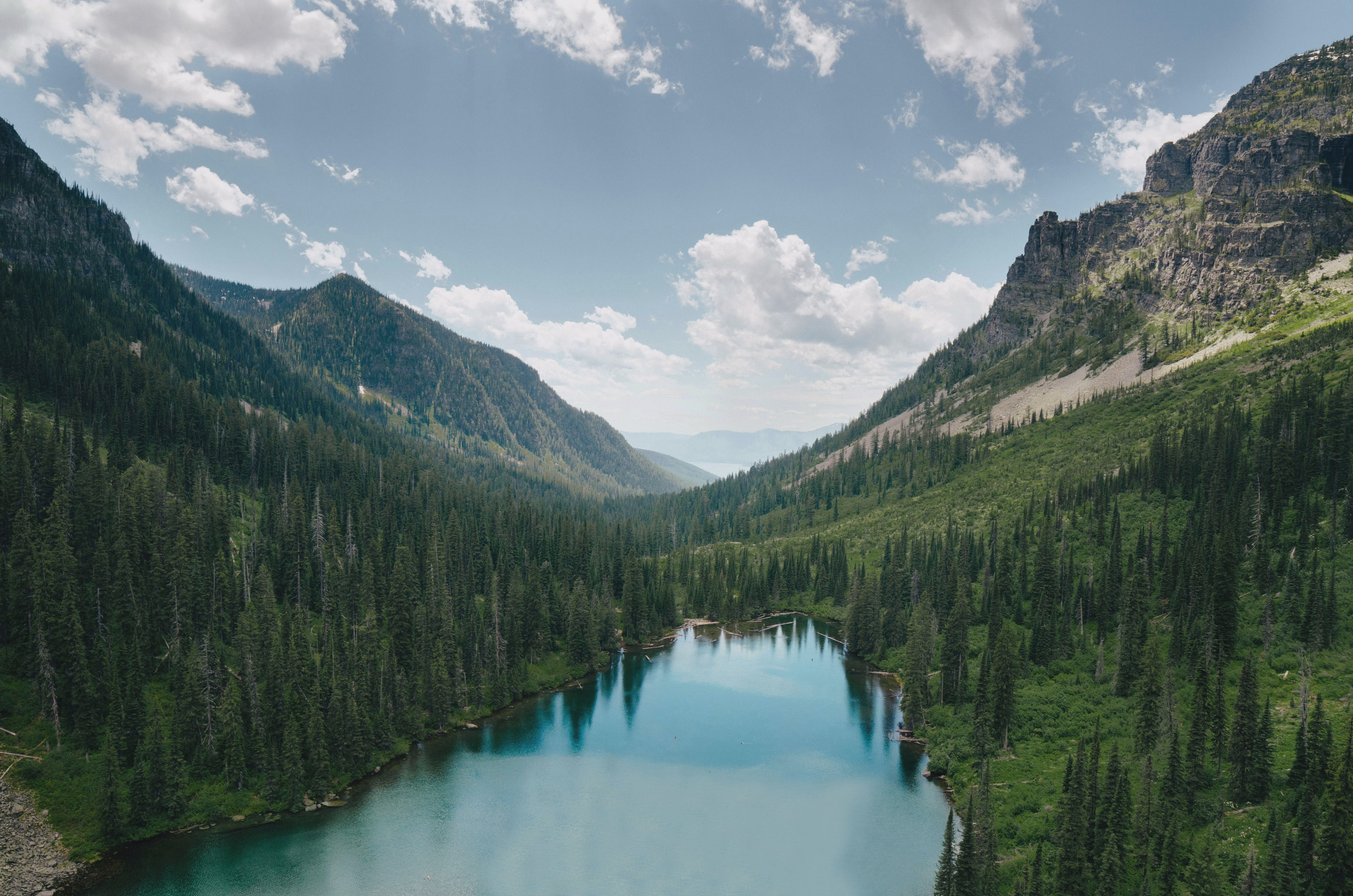 Turquoise alpine lake ringed by dense evergreen forest and steep rocky mountain walls beneath a blue, cloud-dotted sky.