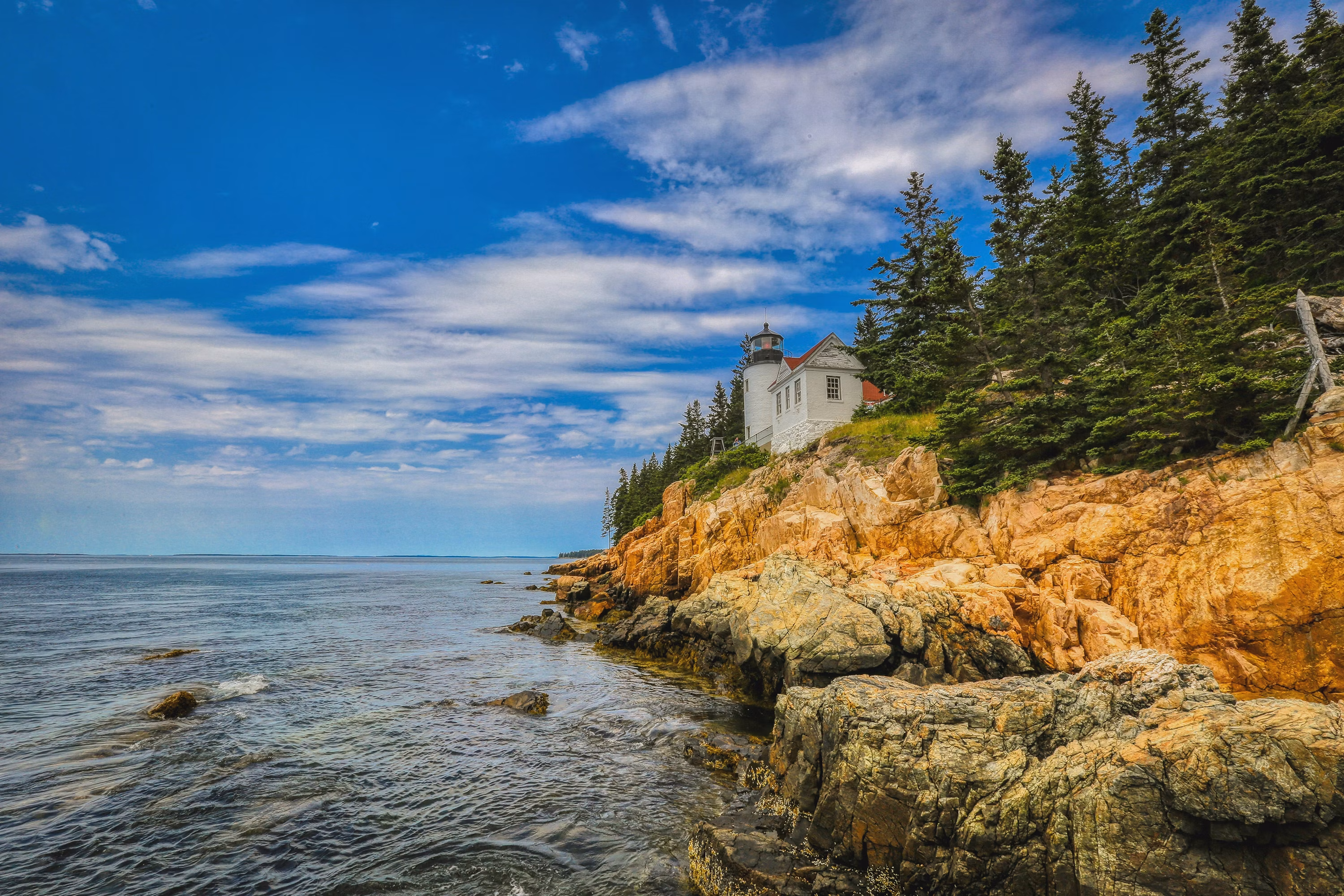 Bass Harbor Head Light stands above a rust-colored granite cliff edged with evergreen trees and blue ocean.