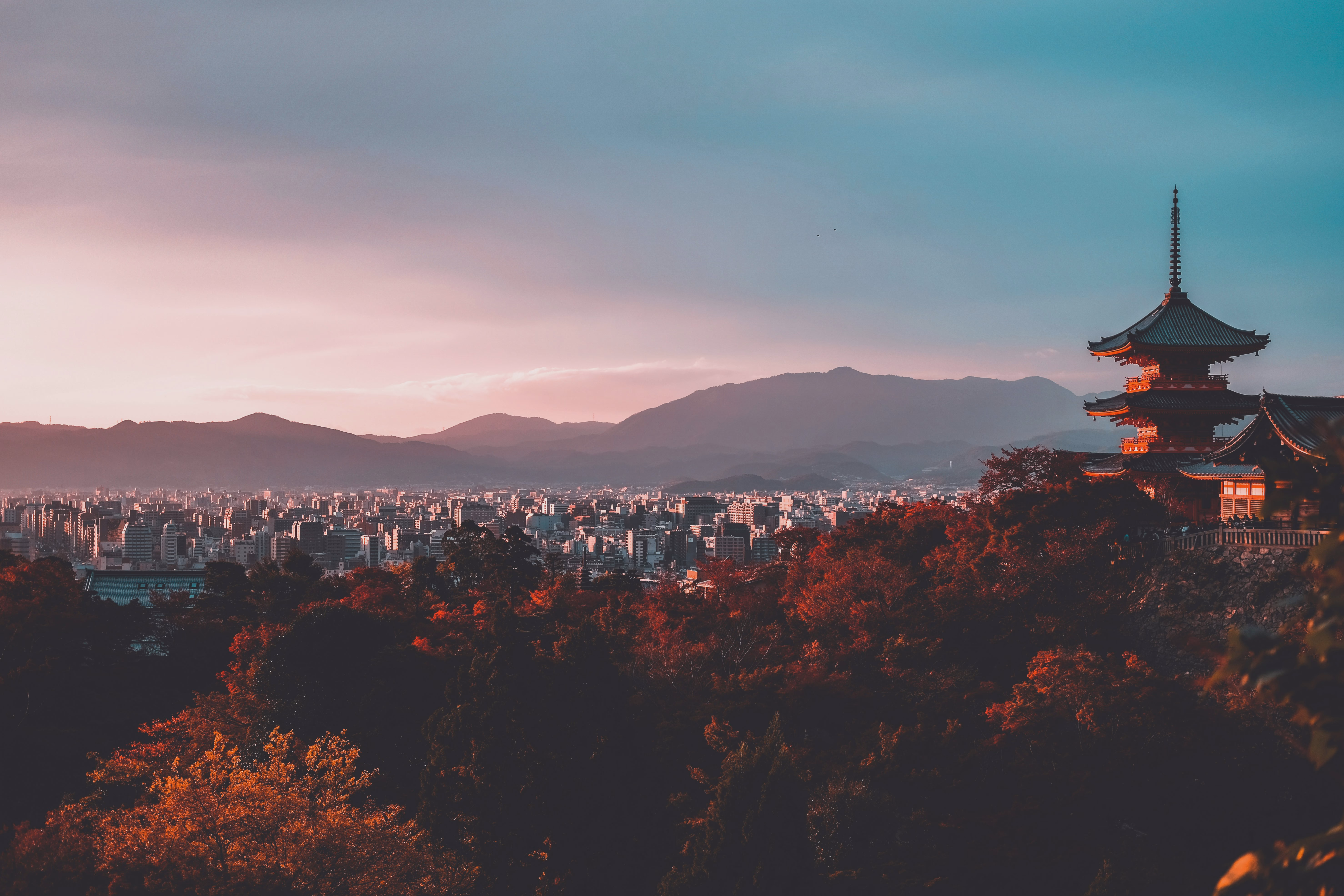 Kyoto's skyline at sunset, with a tiered pagoda silhouetted against hazy mountains and autumn foliage in the foreground.